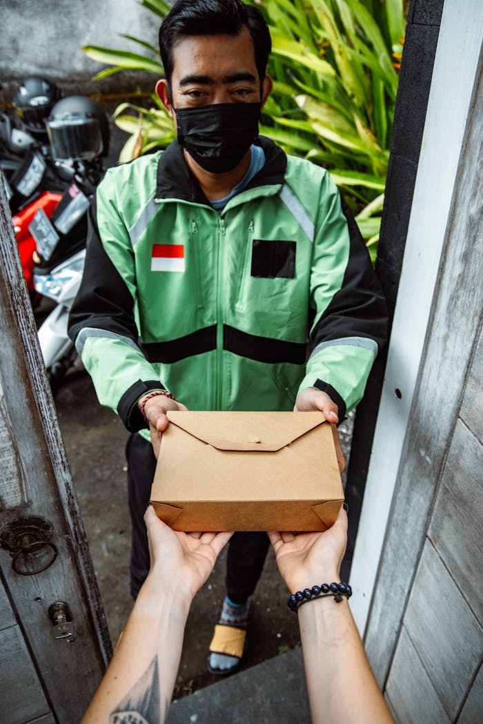 Masked delivery person handing over a food package through a doorway for safe, contactless delivery.